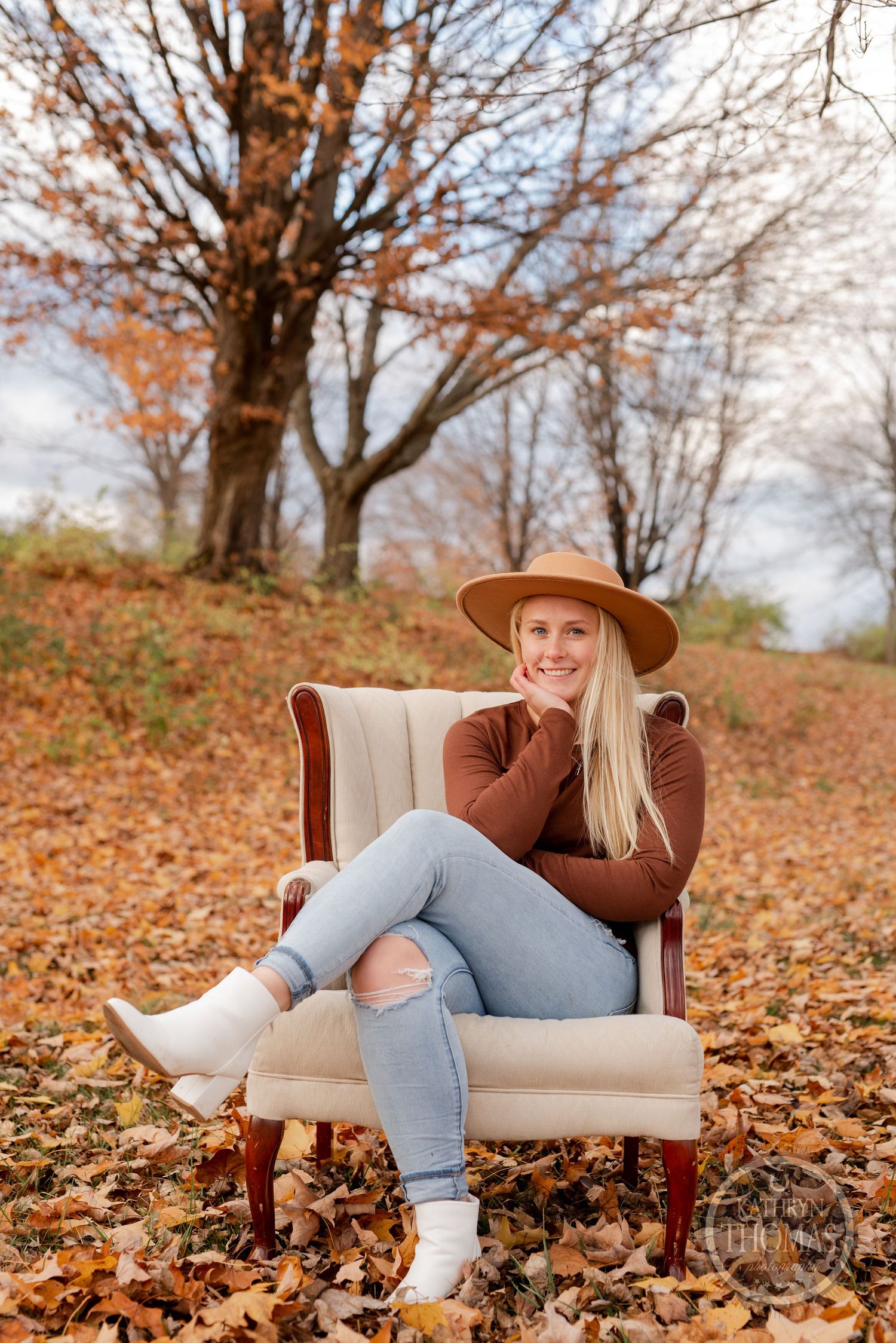 Relaxed autumn portrait in a beige armchair surrounded by fallen leaves and trees.