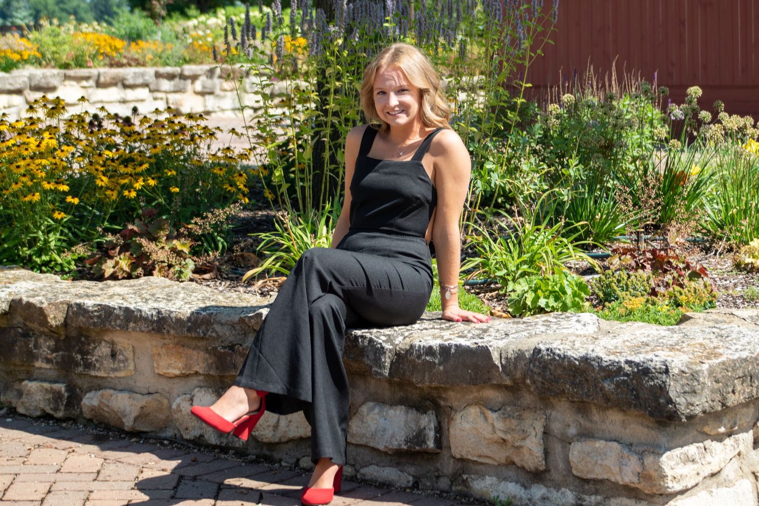 Woman in black dress and red shoes sits on stone wall in garden with yellow flowers.