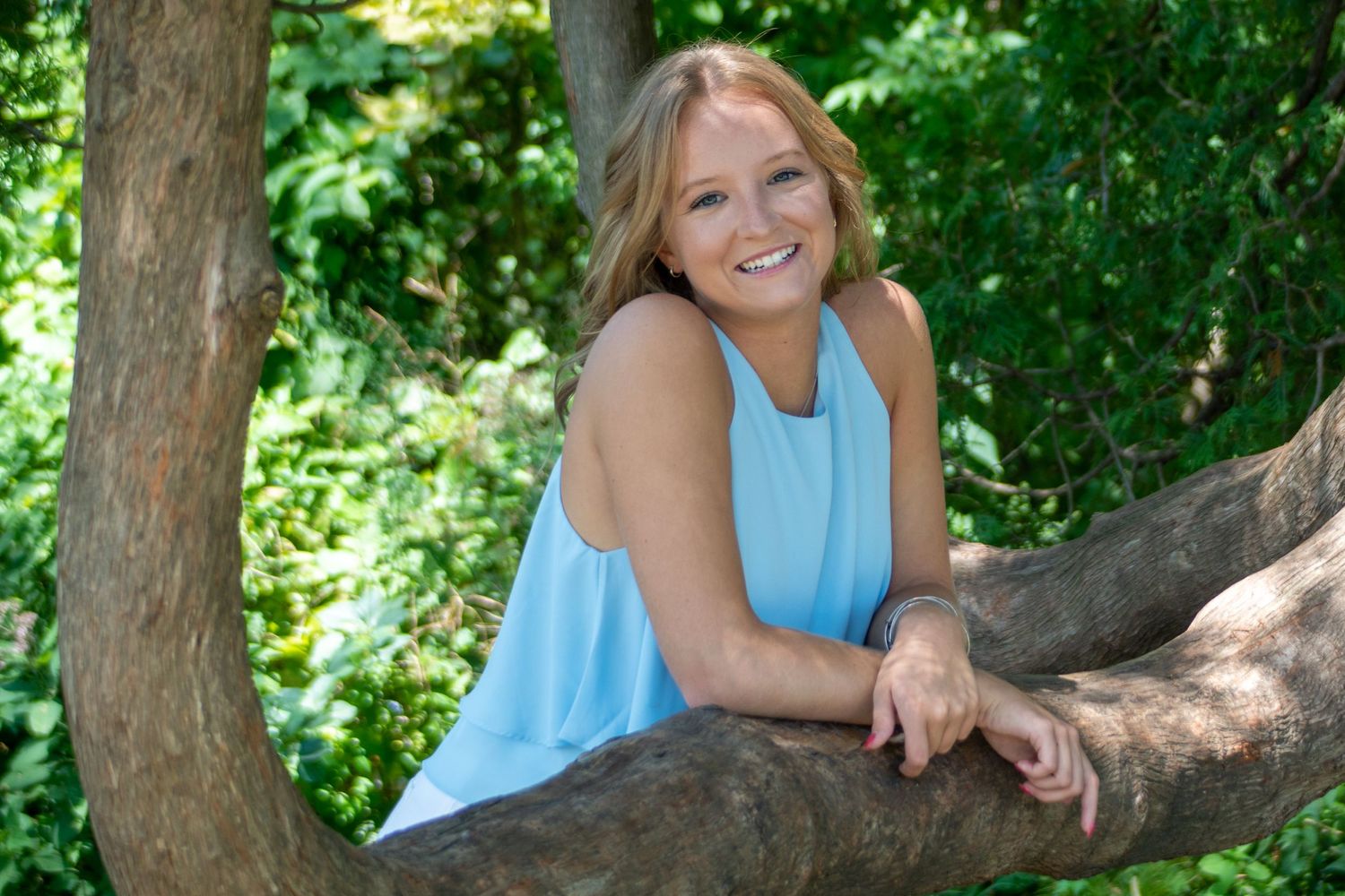 Casual outdoor portrait by tree in dappled sunlight with natural greenery background.