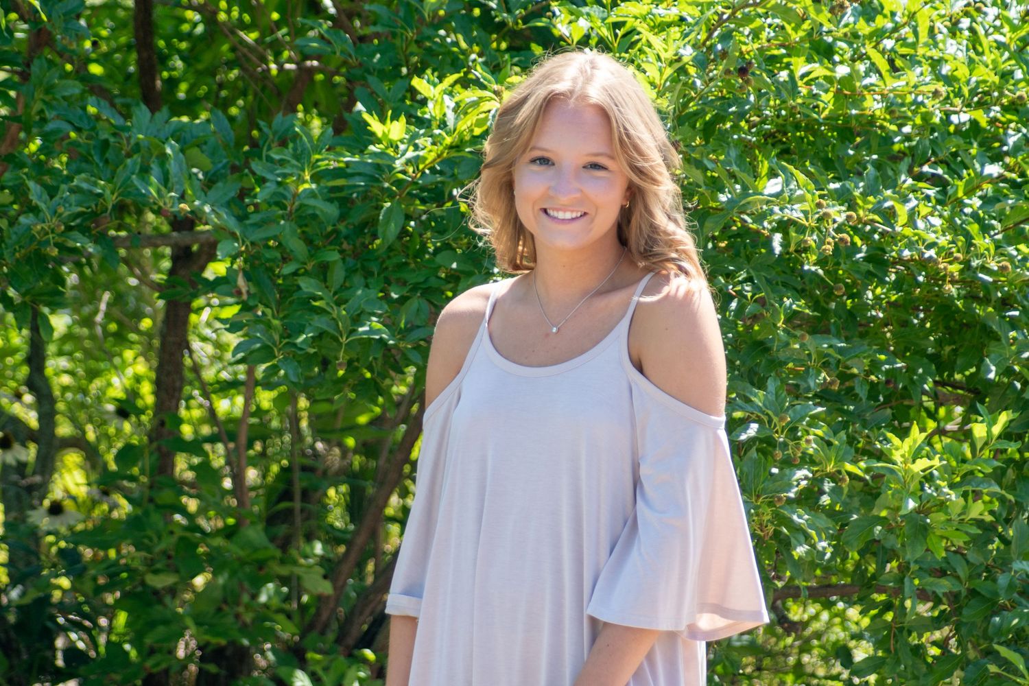A person in a white cold-shoulder dress poses outdoors against a green leafy background.