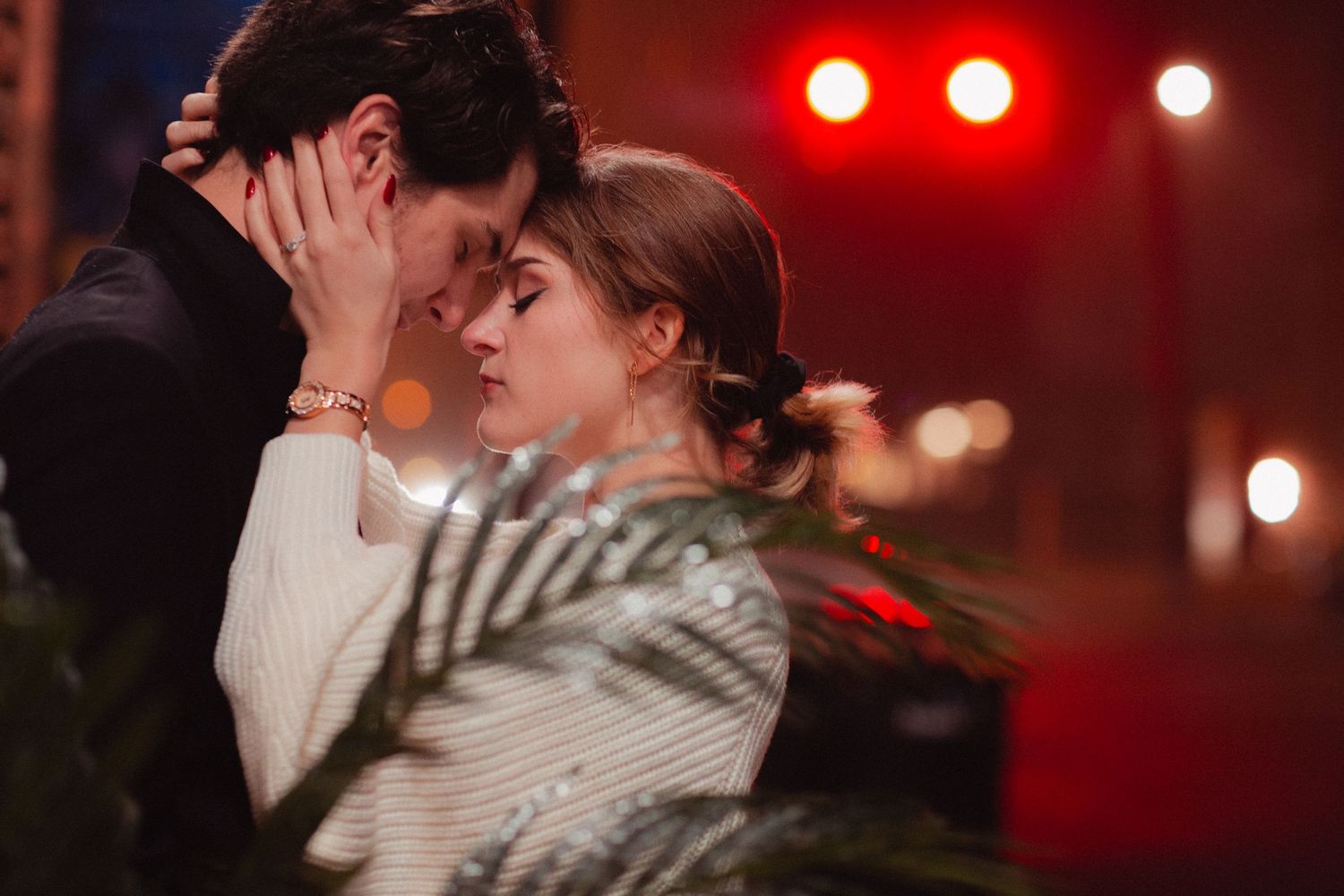 A romantic couple share an intimate moment under red stage lighting with palm fronds in the foreground.
