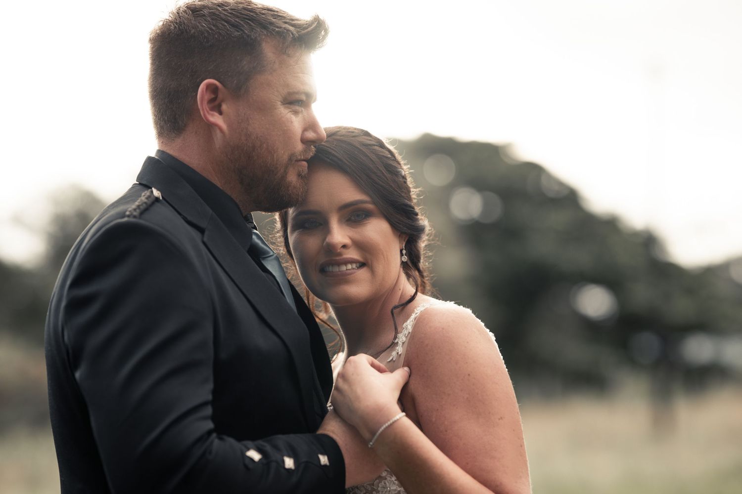 Groom in military dress uniform kissing bride on the cheek in a soft outdoor portrait.