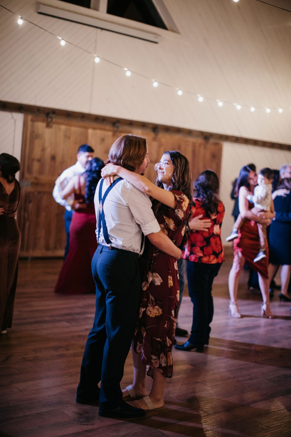 Couple smiling and dancing closely at a wedding reception with string lights and barn doors in background.