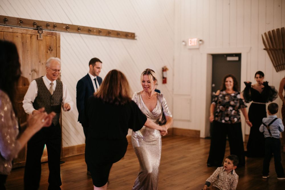Bride in silver gown dancing joyfully with a guest on the wedding reception dance floor in a white barn venue.