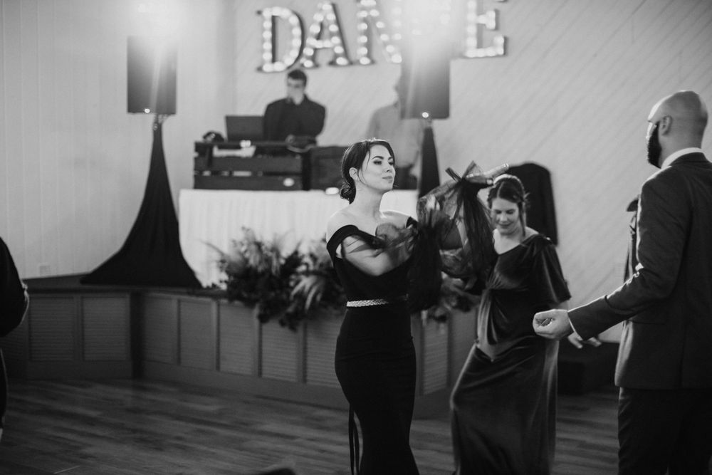 Black and white photo of elegantly dressed wedding guests dancing at a reception with a illuminated DANCE sign backdrop.