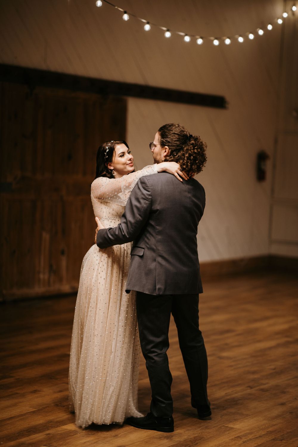 Bride and groom share their first dance in a rustic barn venue adorned with warm string lights overhead.