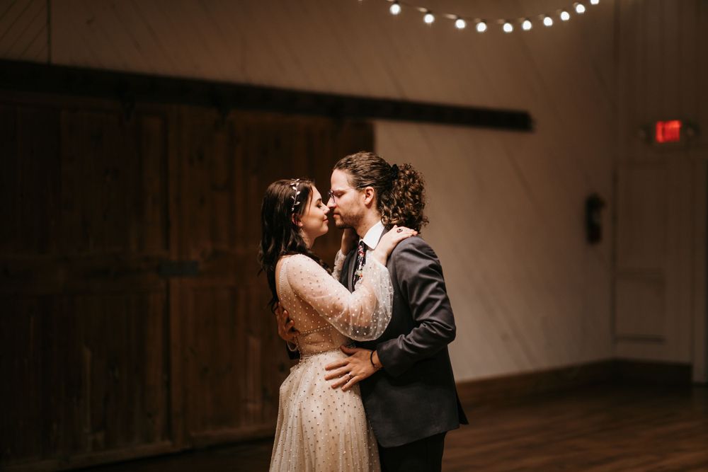 Bride and groom share a romantic kiss during first dance in warmly lit rustic barn wedding venue.