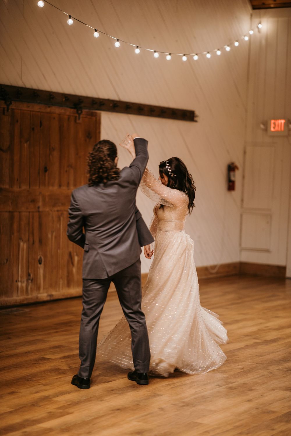 Groom twirls bride in sparkling wedding gown during first dance in elegant barn venue with string lights.