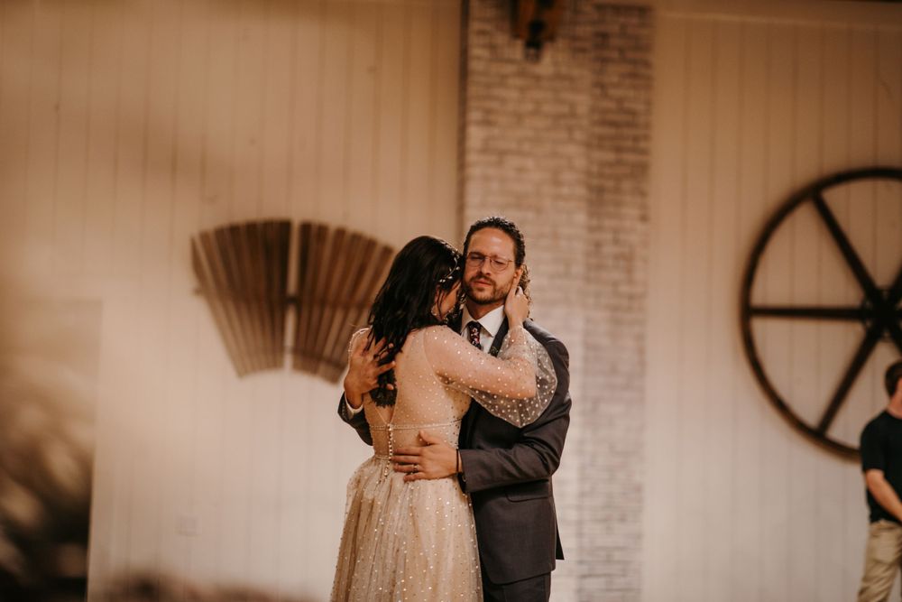 Bride and groom share a tender embrace during first dance in warmly lit rustic wedding barn venue.