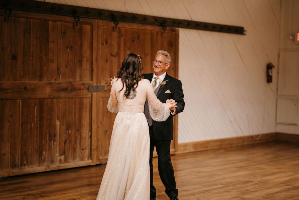 Bride shares a joyful father-daughter dance in elegant rustic barn wedding venue with wooden barn doors.