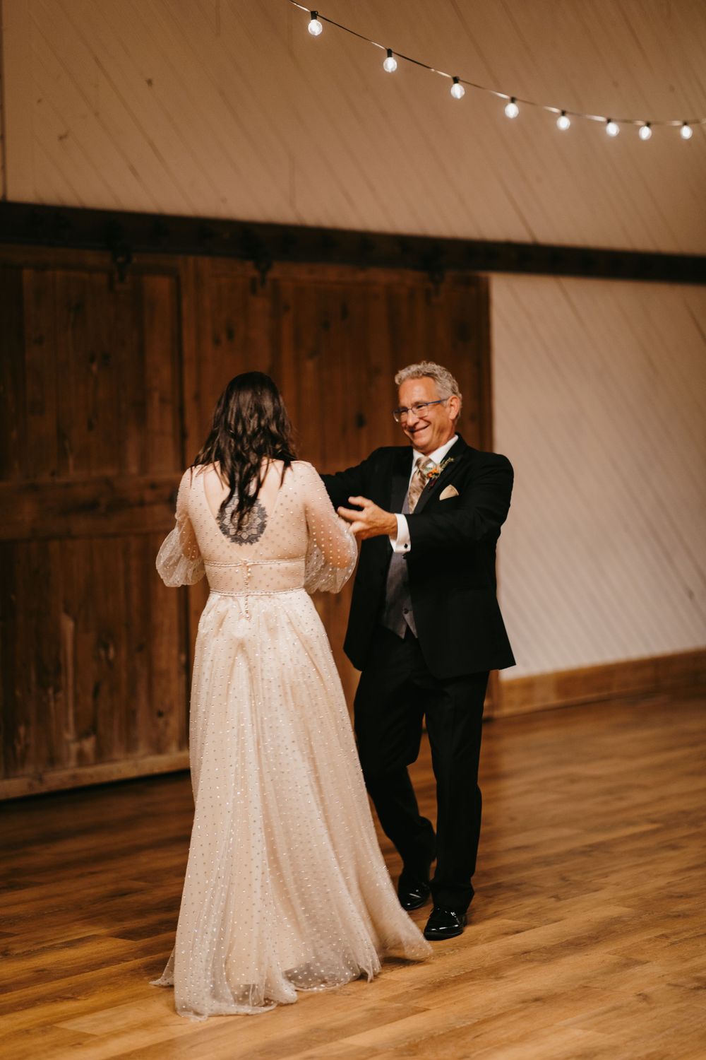 Bride dances with her smiling father in a rustic barn venue decorated with overhead string lights.