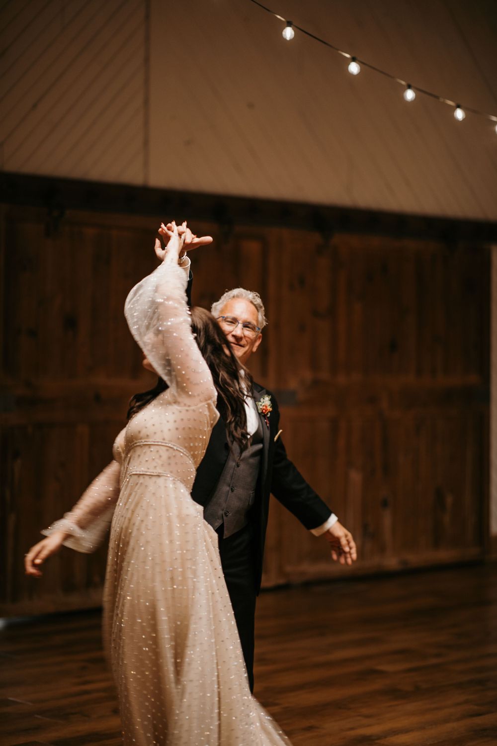 Bride and older gentleman share a joyful first dance in a rustic barn venue with string lights overhead.