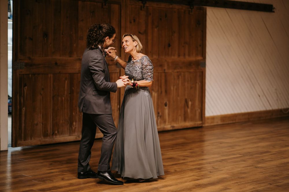 Groom and elegant woman in grey gown share a graceful dance in front of rustic wooden barn doors.