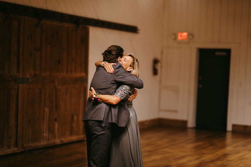 Two people share an emotional hug on the wooden dance floor of a rustic barn wedding venue.