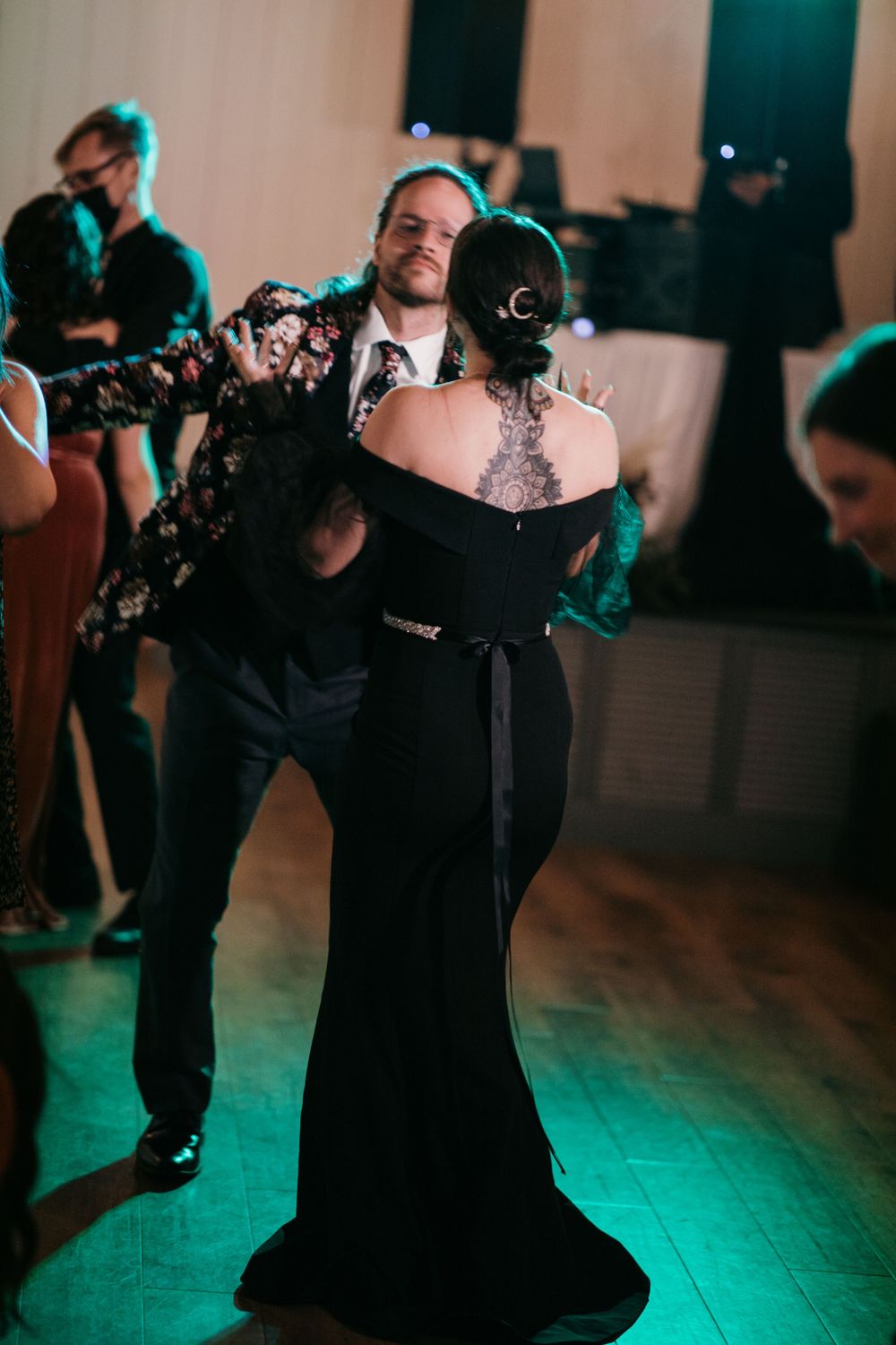 Bride in elegant black off-shoulder gown with back tattoo shares first dance with groom in floral jacket.