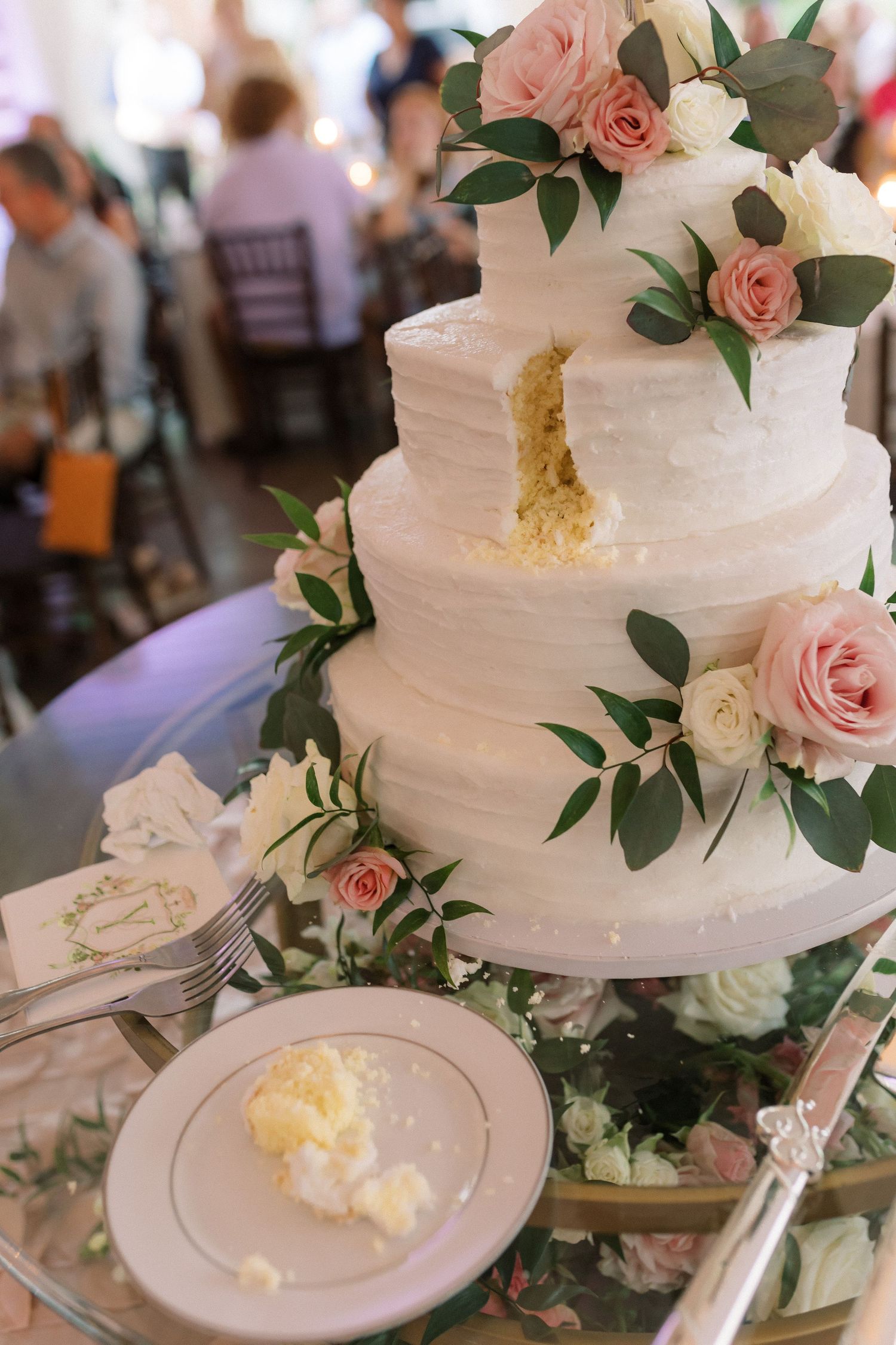 An elegant white wedding cake decorated with pink roses and greenery sits on a decorated table.