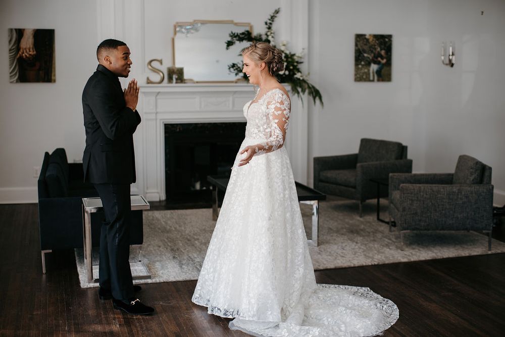 Groom in black suit clasps hands in awe seeing bride in lace wedding gown during first look indoors.