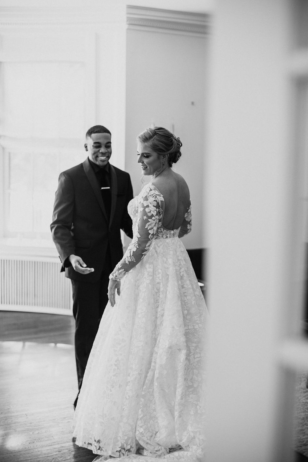 Black and white photo of smiling bride in lace gown and groom in black suit sharing joyful moment.