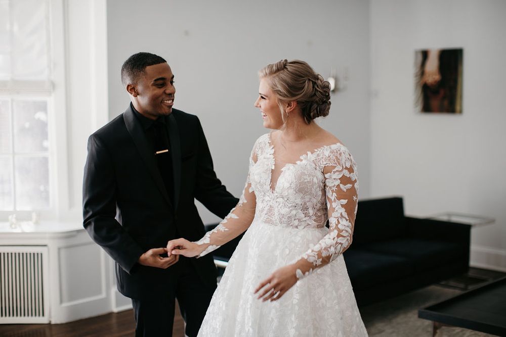 Groom in black suit holds bride's hands admiring her floral lace long-sleeve wedding gown indoors.