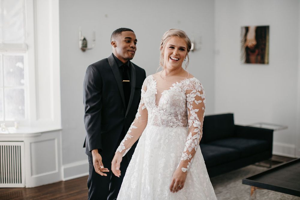 Groom stands behind beaming bride in lace wedding dress during romantic indoor first look reveal.