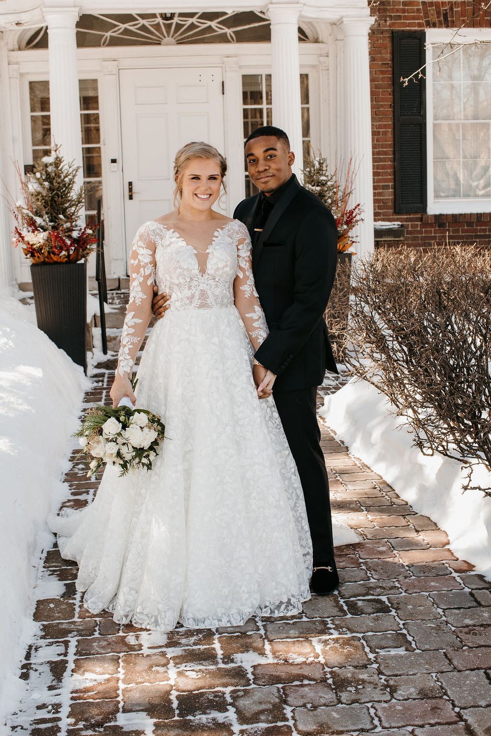 Bride in lace long-sleeve gown and groom in black suit pose on snowy brick pathway outdoors.
