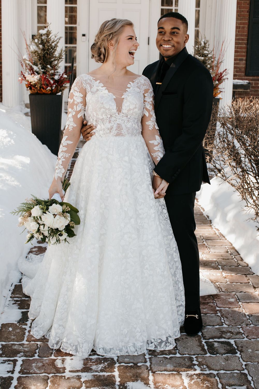 Laughing bride holding white bouquet and grinning groom in black suit pose in snowy winter setting.