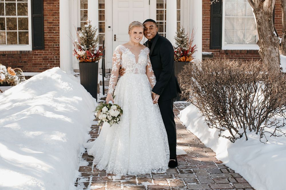 Bride in lace gown holding white bouquet and groom in black suit pose in snowy outdoor setting.