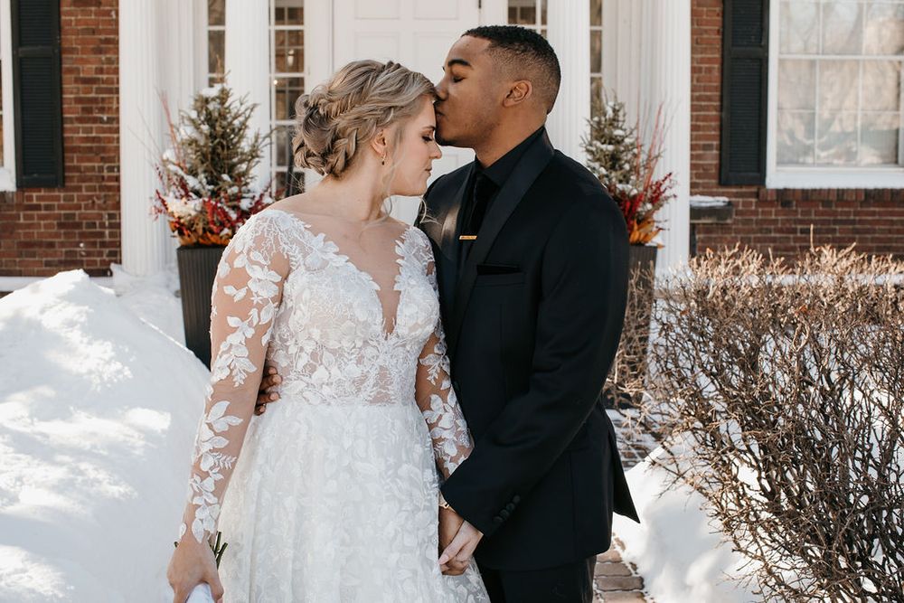 Bride and groom share a romantic kiss outside a brick building surrounded by winter snow.