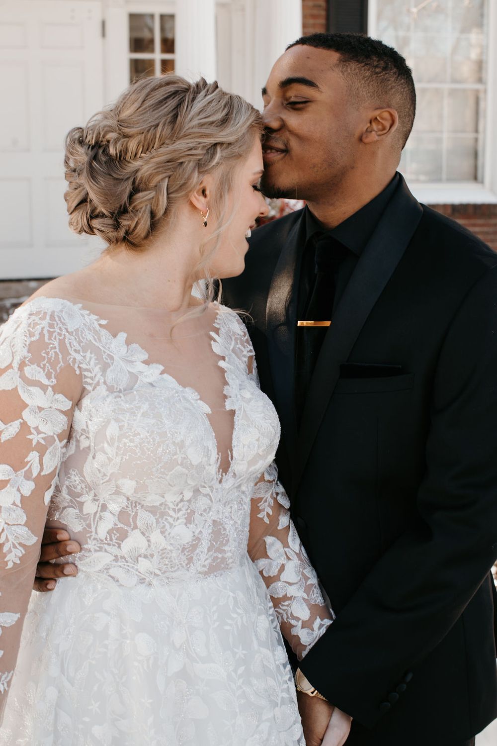 Interracial couple sharing a tender moment, groom in black suit kissing bride wearing lace long-sleeve wedding gown.
