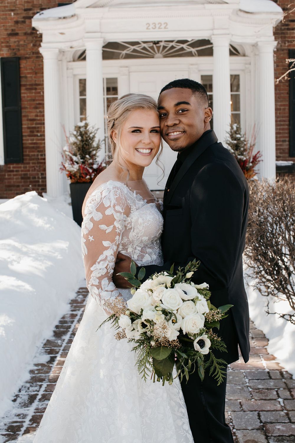 Smiling interracial wedding couple posing in snow outside brick building, bride holding white floral bouquet.