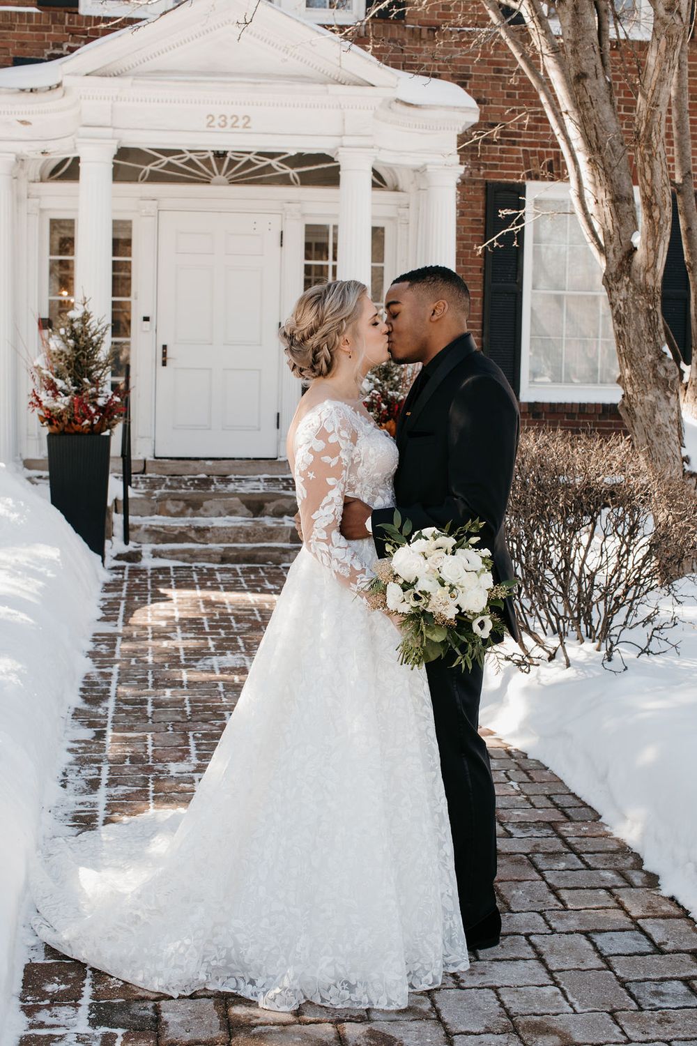 Interracial bride and groom sharing a kiss on snowy brick pathway outside elegant building, bride holding bouquet.