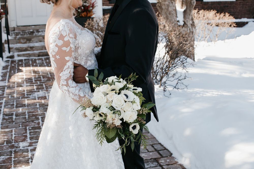 Close-up of bride in lace wedding gown and groom holding white and green bridal bouquet in snowy outdoor setting.