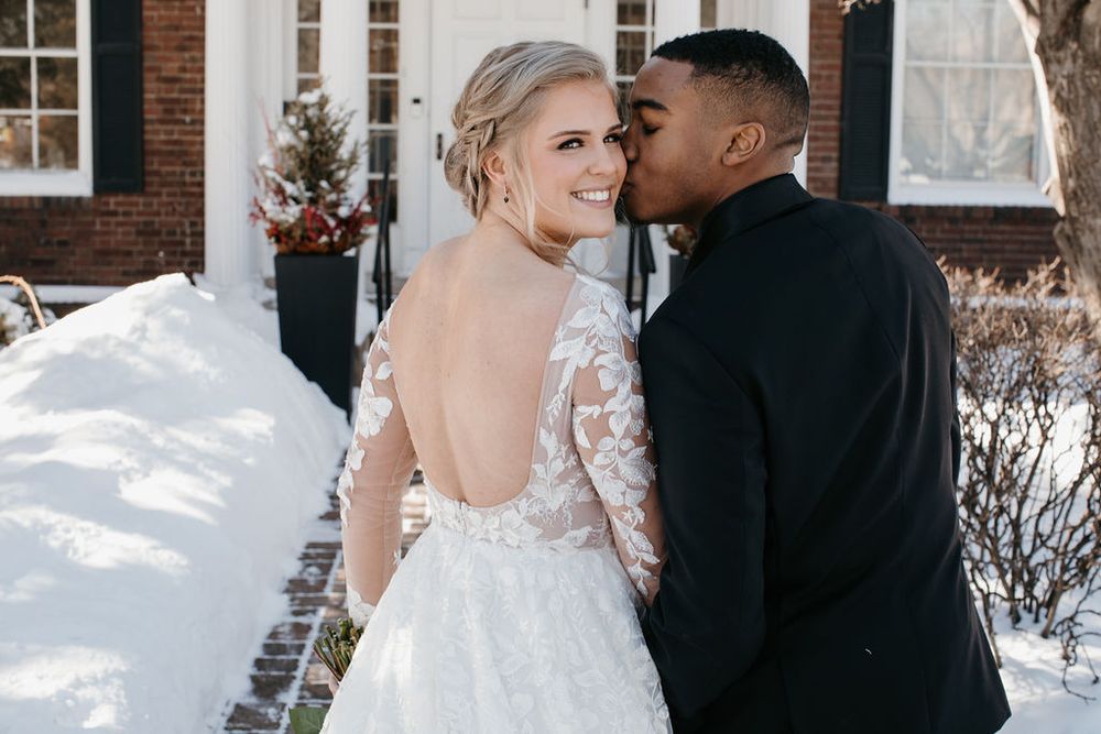 Groom in black suit kissing bride on cheek, bride smiling in backless lace wedding gown in winter setting.