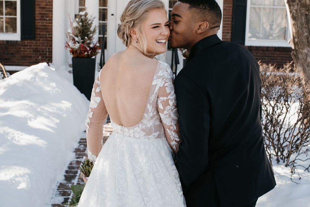 Groom kissing smiling bride on cheek outdoors in snow, bride wearing elegant backless lace long-sleeve wedding dress.