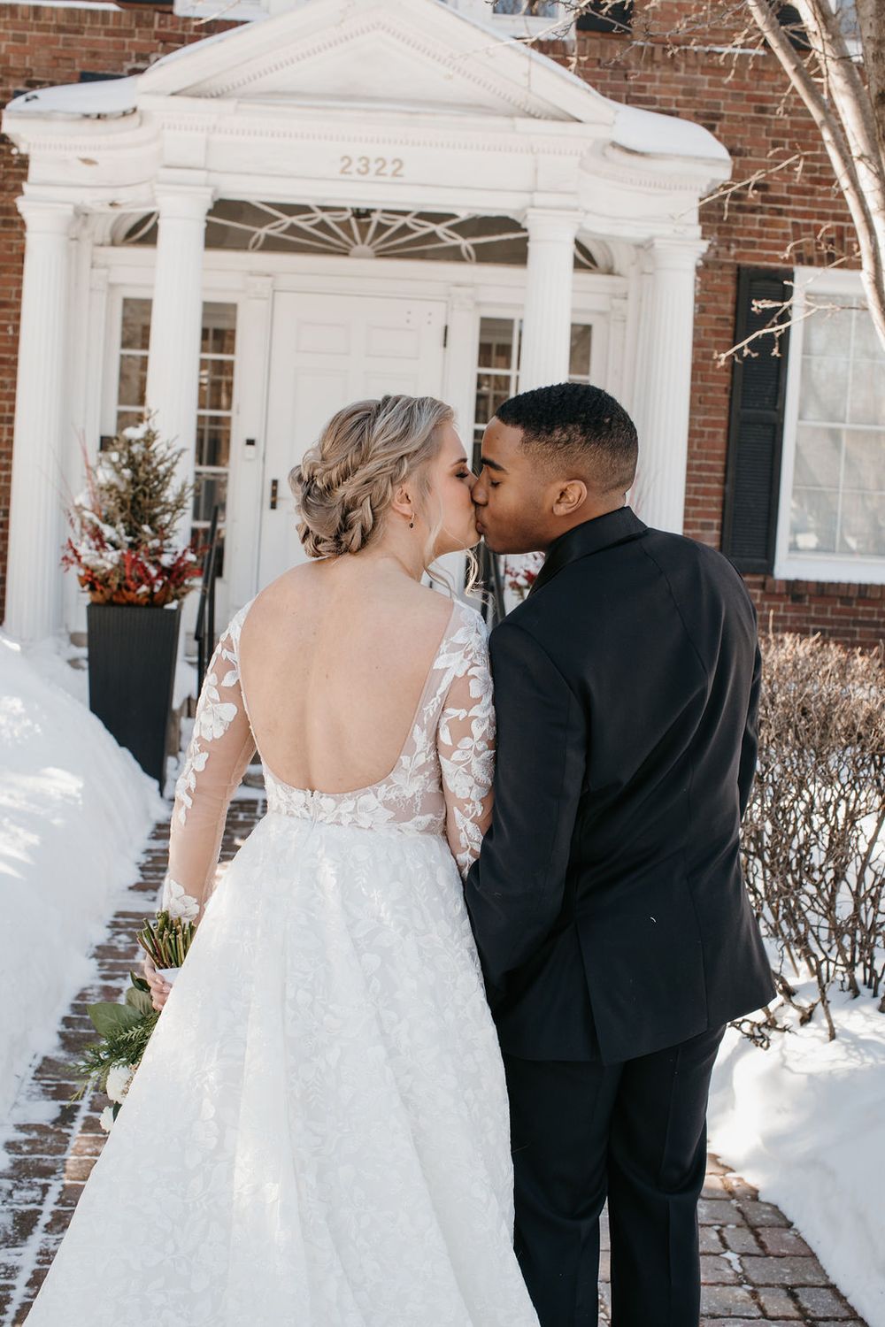 Interracial couple kissing outside brick building numbered 2322 in winter, bride in backless lace gown.