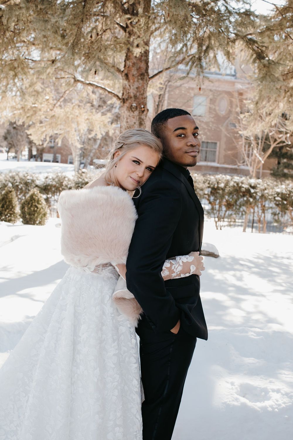 Interracial bride and groom posing back-to-back under snow-covered tree, bride in lace gown with blush fur wrap.
