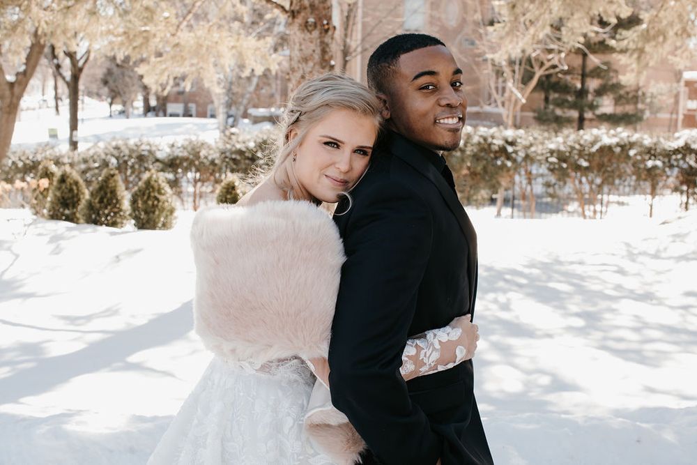 Interracial wedding couple standing back-to-back in snowy park, bride wearing white fur wrap over lace gown.