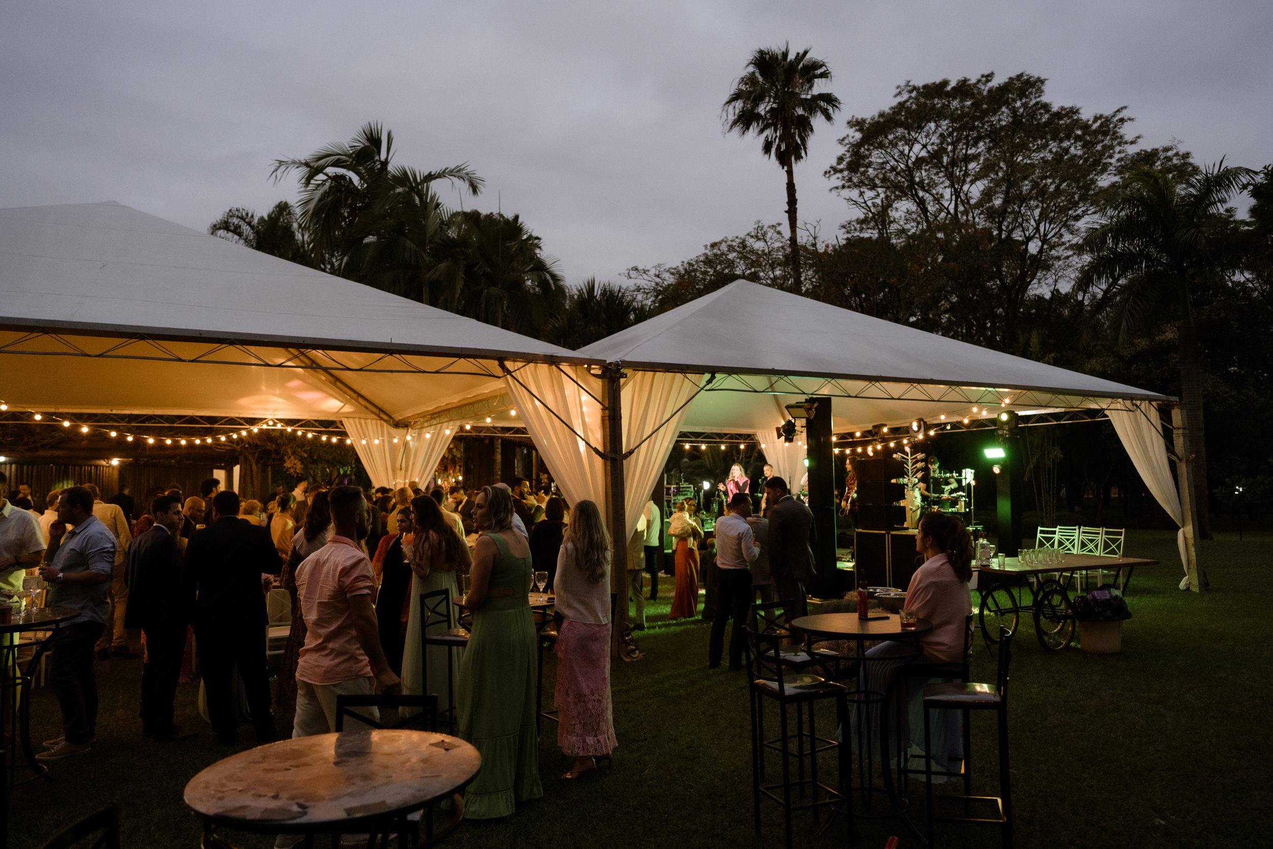 Evening wedding reception under illuminated white tents with guests mingling on a lawn.