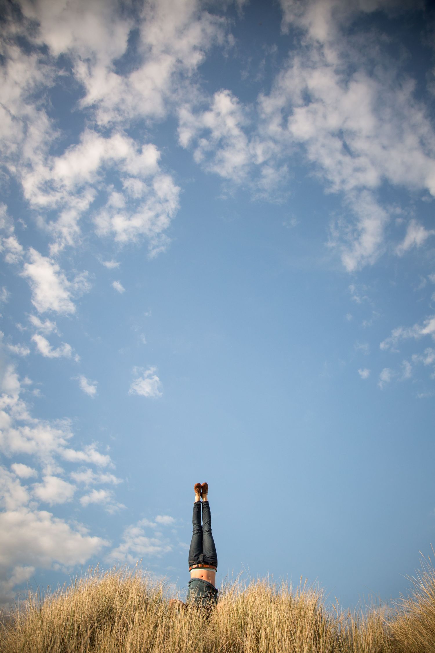 Person doing handstand against blue sky with white clouds on grassy hill.