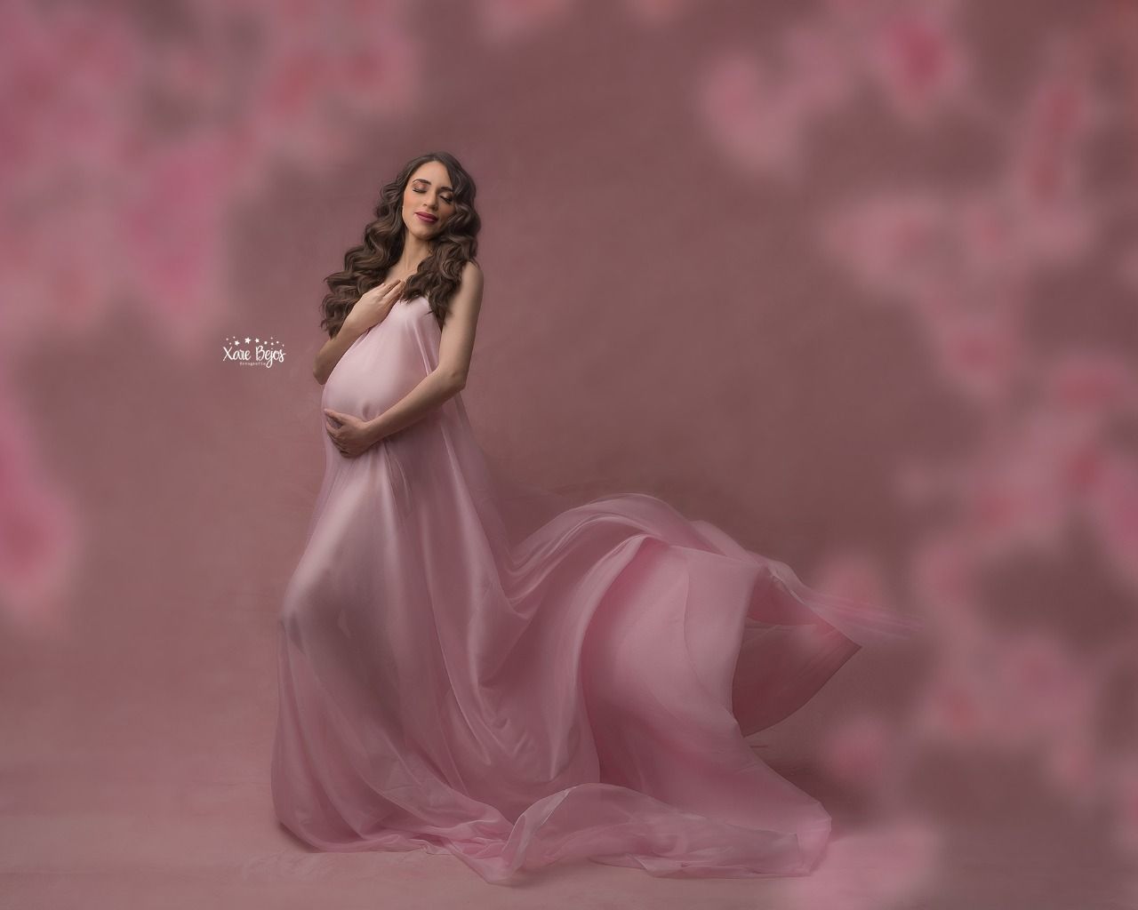 Dreamy maternity portrait in flowing pink gown against rosy background.