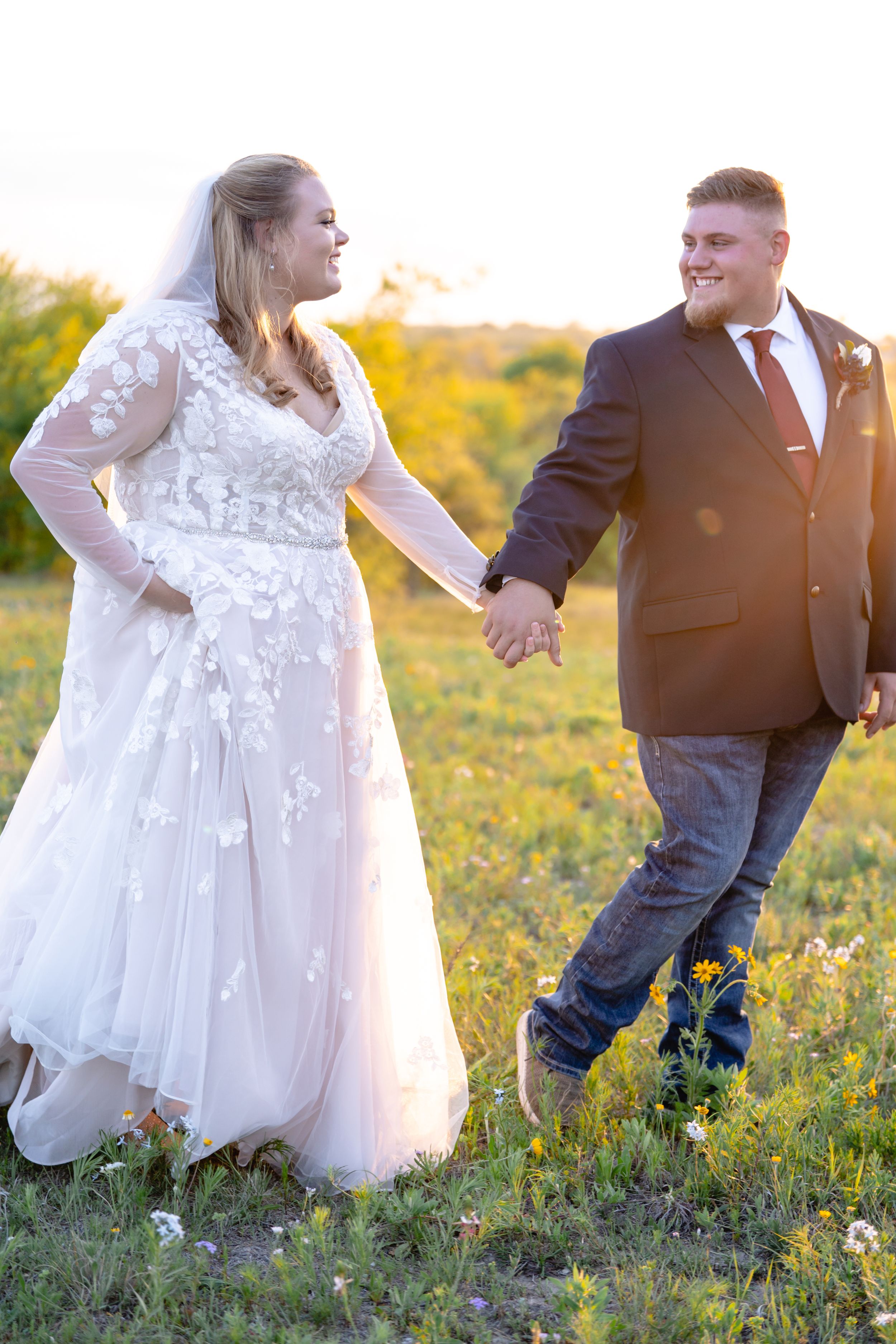 Newlyweds posing playfully in a sunlit field with bride in lace dress and groom in dark suit.