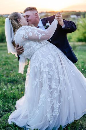 Wedding couple dancing and embracing in a meadow at sunset with bride in flowing lace gown.