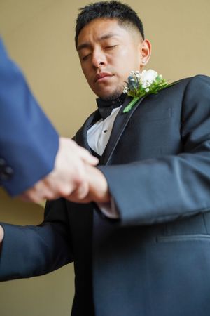 Groom in black tuxedo having his boutonniere pinned on during wedding preparations.