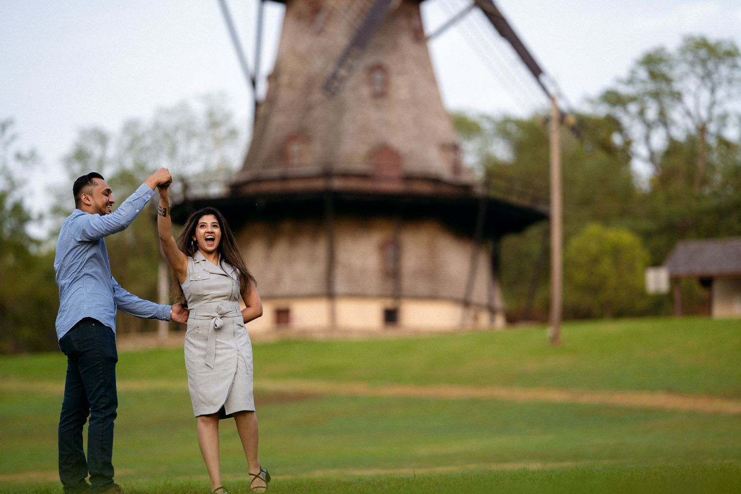 Groom spins bride in front of vintage windmill, verdant lawn, encapsulating joy and timeless romance in their engagement