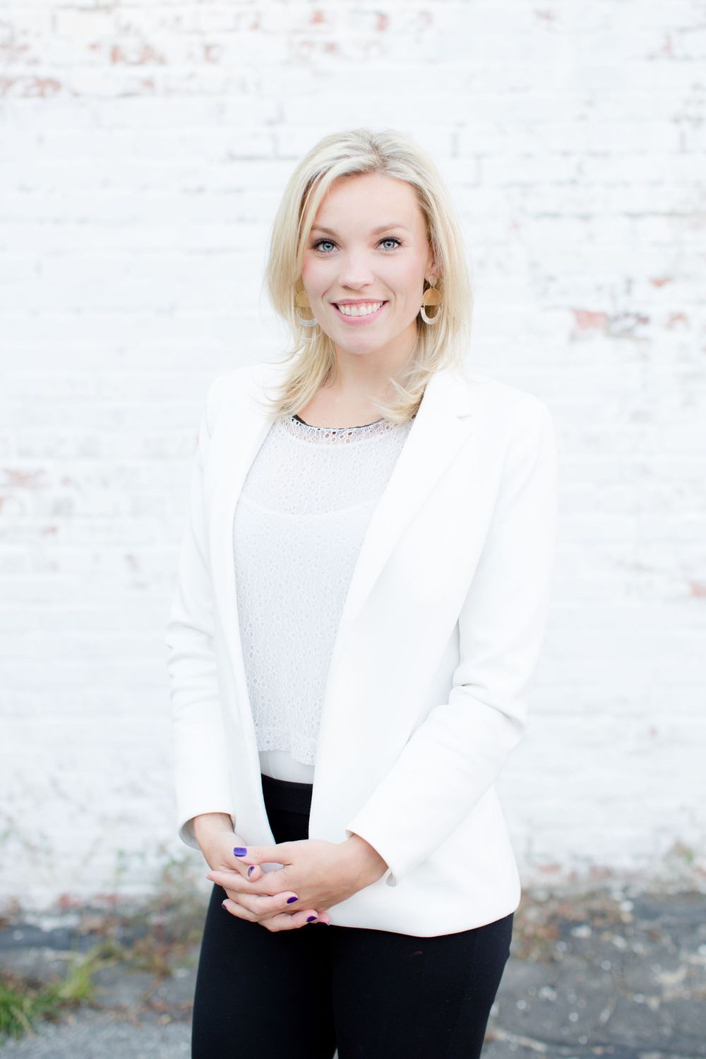 Professional headshot of woman in white blazer against brick wall.