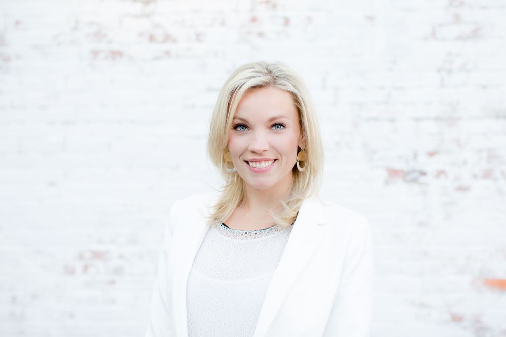 Portrait of woman in white blazer smiling against weathered wall background.