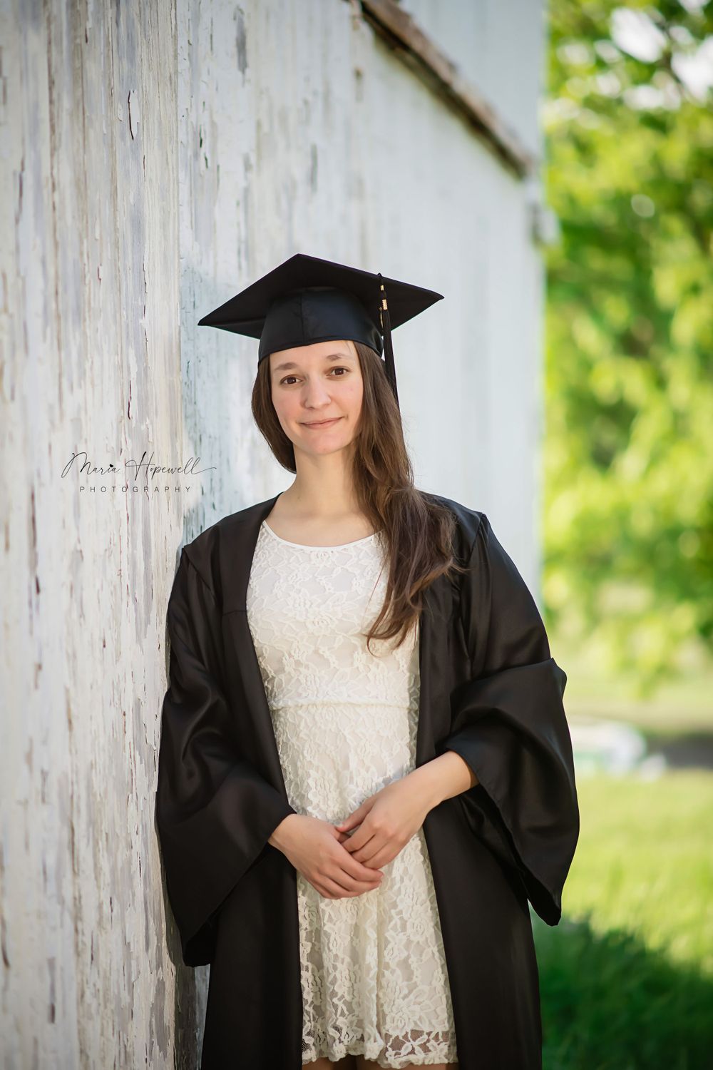 Graduate in black cap and gown with white lace dress standing against rustic wall.