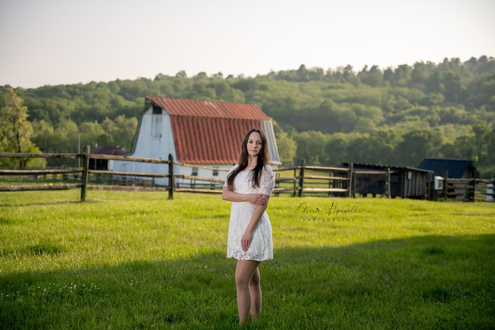 Woman in white dress posing in green field with rustic barn in background.