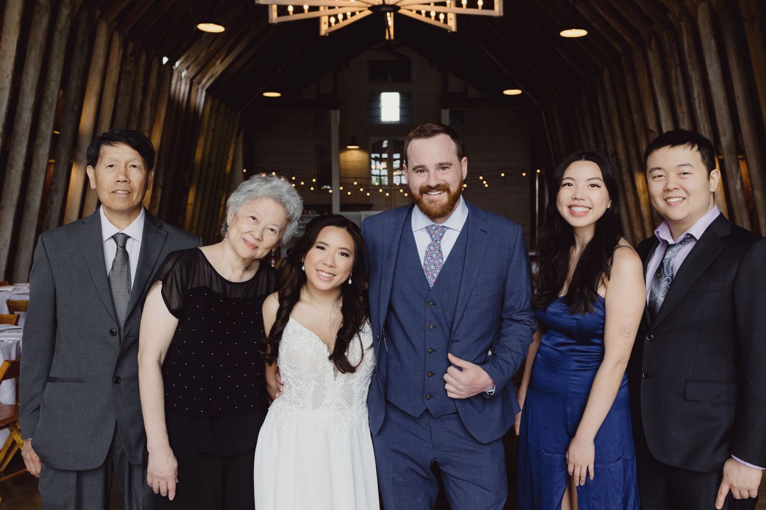 A wedding party group poses together inside a dark wooden barn-style venue.