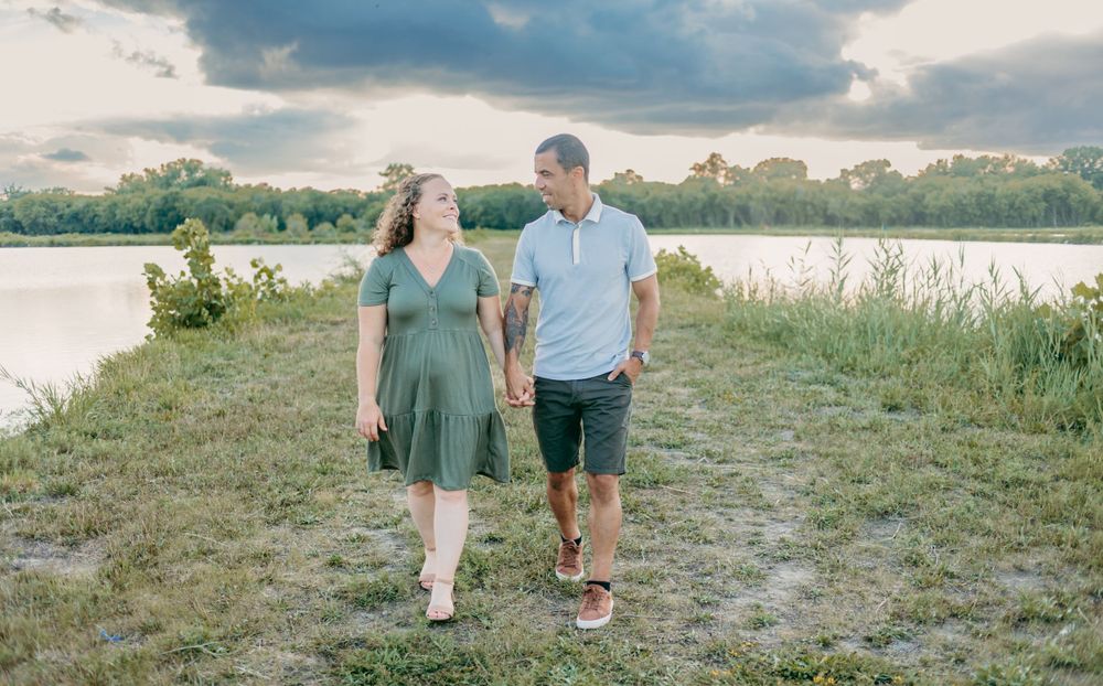 Couple walks together on grassy path near water during golden hour sunset.